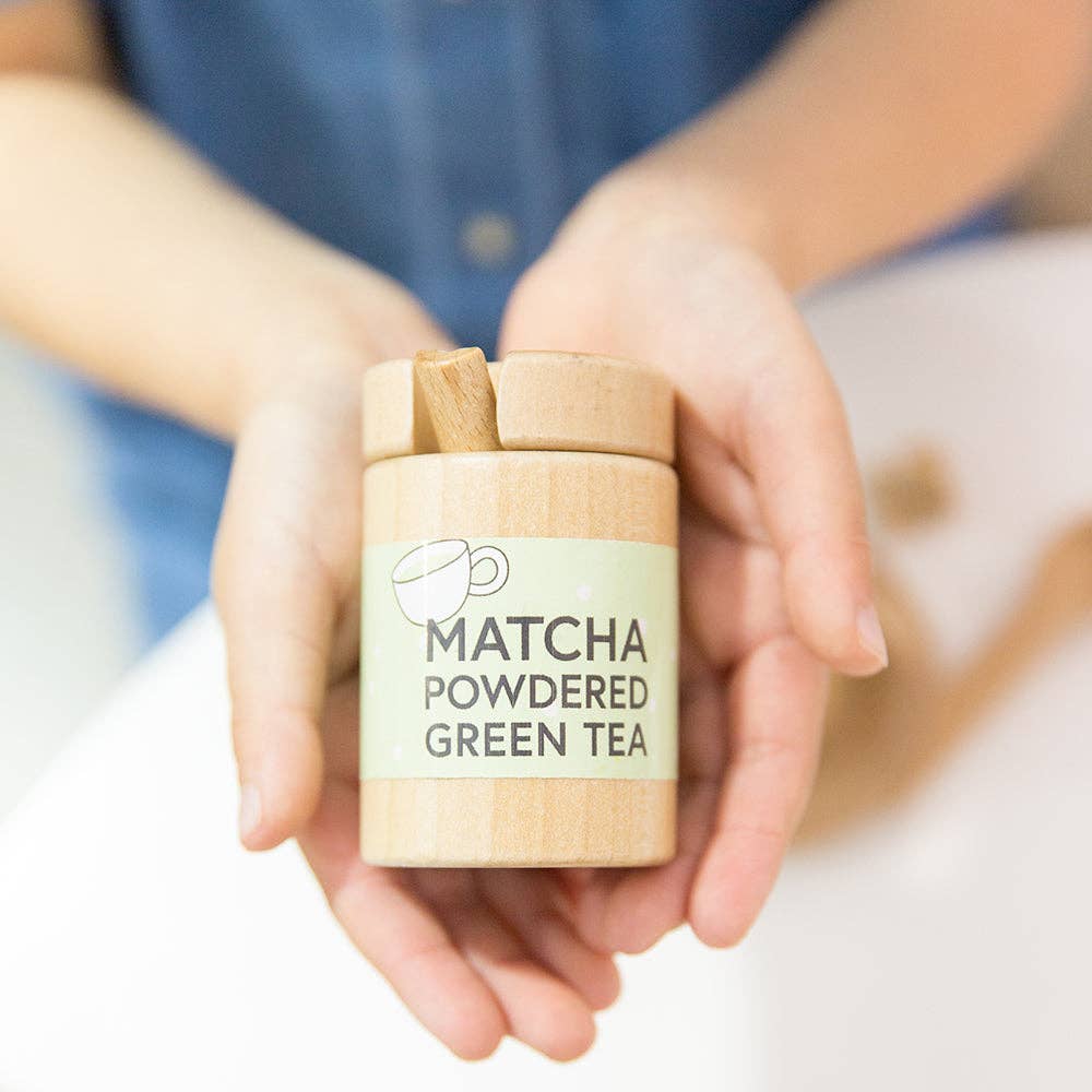 Hands cradle a small wooden tea set labeled "Organic Matcha Powdered Green Tea," adorned with a teacup image. A wooden spoon rests atop, enhancing the scene's natural elegance against a softly blurred background.