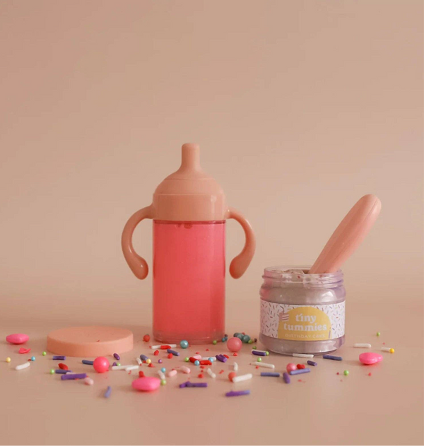 A pink magical strawberry milk cup with handles is next to an open "tiny tummies" jar filled with creamy goodness, surrounded by colorful sprinkles and small candies, creating a whimsical doll's party scene. This enchanting setup feels straight out of the My Tiny Birthday Set - Tiny Harlow on a plain backdrop.