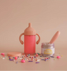 A pink magical strawberry milk cup with handles is next to an open "tiny tummies" jar filled with creamy goodness, surrounded by colorful sprinkles and small candies, creating a whimsical doll's party scene. This enchanting setup feels straight out of the My Tiny Birthday Set - Tiny Harlow on a plain backdrop.