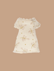 The Maileg Little Sister Bedtime Set features a small, cream-colored dress with short sleeves and pink floral patterns, perfect for Maileg dolls. Displayed on a solid beige background.