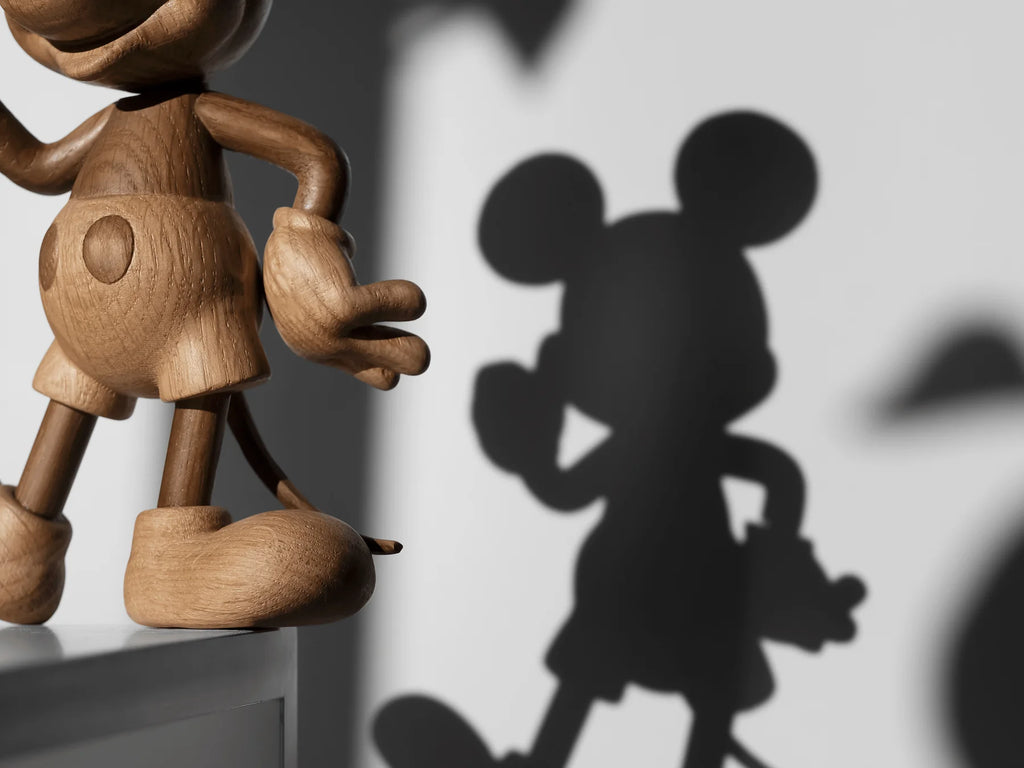 A wooden figure named Mickey Mouse, Oak is partially visible on the left, casting a shadow on a gray wall. The shadow reveals Mickey Mouse’s iconic round ears and gloves, capturing the adventurous spirit of this global icon.