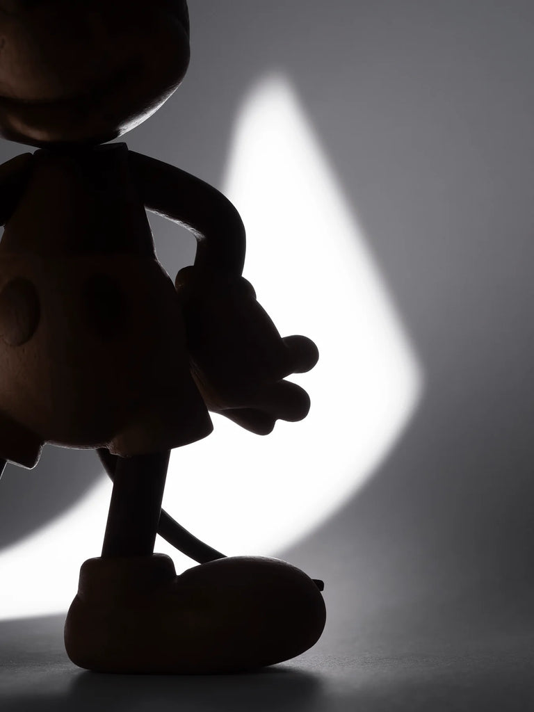 Silhouette of a Mickey Mouse, Oak figurine stands against a bright, soft-focus, light gray background. Partially visible, it echoes Mickey's adventurous spirit with its large round shoes and gloved hands.