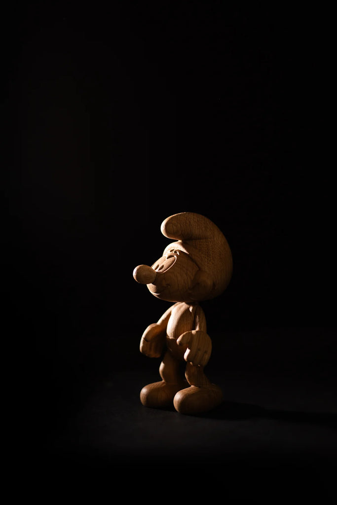 The Smurf, Oak figurine, a small wooden character resembling those from Smurf Village, stands illuminated against a dark background. Its hat and large shoes cast a subtle shadow that hints at Gargamel lurking nearby.