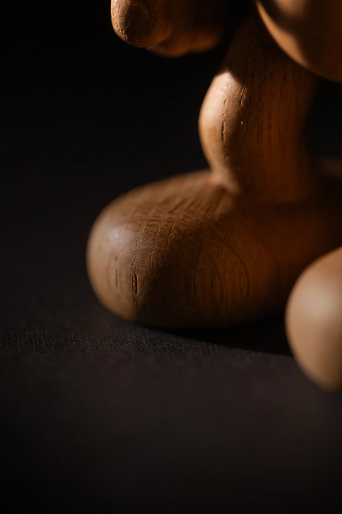 A close-up of the "Smurf, Oak" sculpture highlights its elegantly carved foot and leg under soft lighting, revealing rich wood grain and texture. The scene evokes a mystical aura, as if paying homage to a hidden corner of Smurf Village against a mysterious dark background.