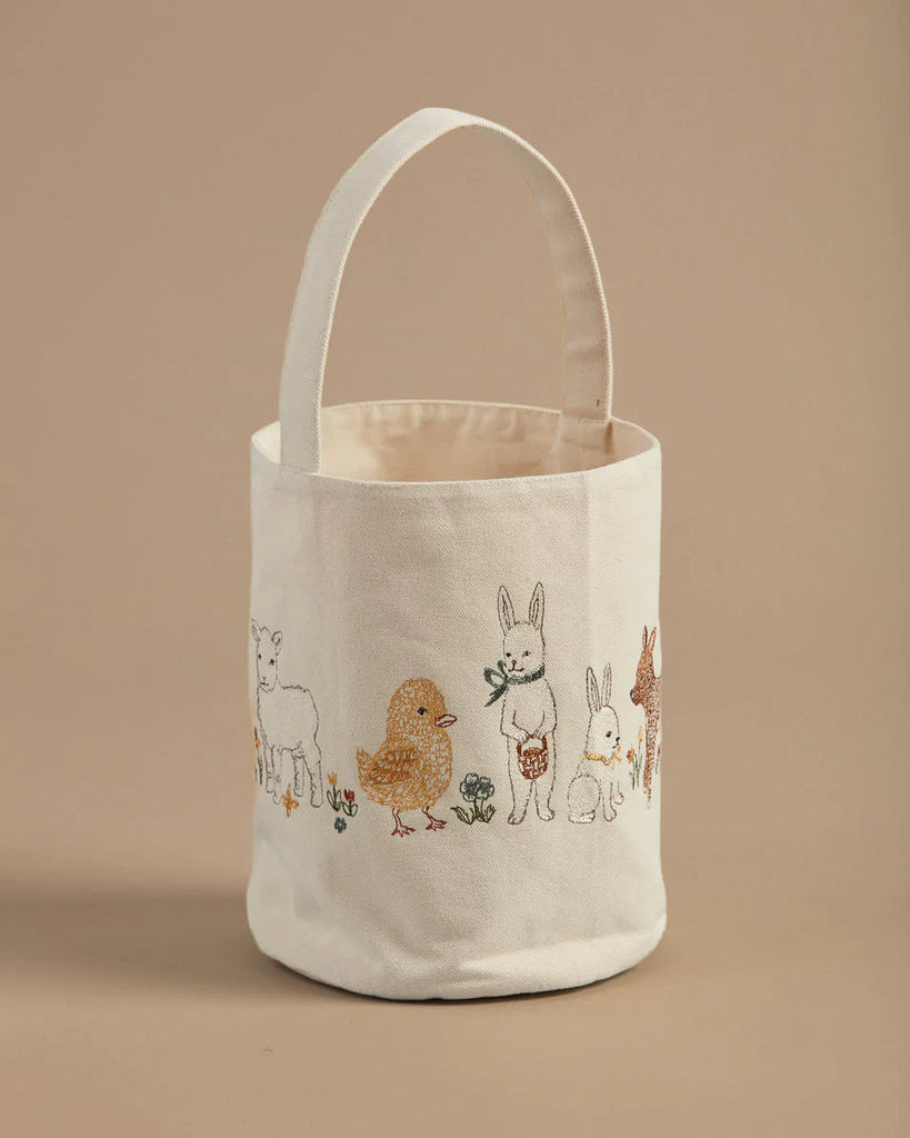 The Coral & Tusk Spring Babies Canvas Bucket is a beige fabric tote with a single handle, designed like an Easter basket. It features embroidered images of a lamb, chick, rabbit, other animals, and small plants on a plain light brown background.
