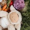 An Olli Ella Cozy Dinkum Doll - Lamby Picket with closed eyes is nestled among fresh vegetables and pink hydrangeas. The setting, with carrots and leafy greens, offers a perfect Easter gift for loved ones.