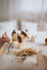 A Handmade Wooden Donkey, crafted from linden wood and painted with child-safe paint, is displayed alongside other farm animals on a textured white surface with a small straw pile. The softly blurred background enhances the cozy, rustic atmosphere.