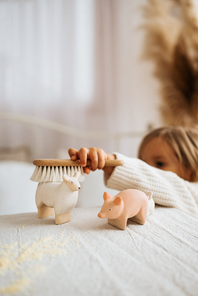 A child in a white sweater plays with a Handmade Wooden Sheep crafted from linden wood on a textured white surface. Soft, neutral tones and pampas grass in the background create a cozy atmosphere, ensuring charming and safe playtime with child-safe paint.