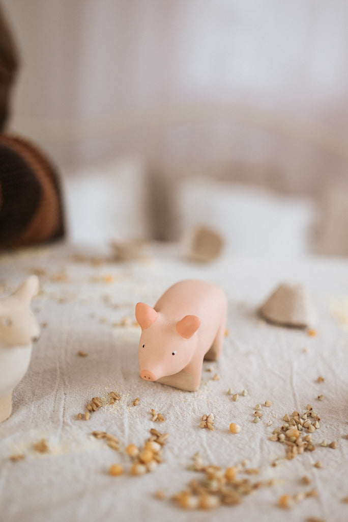 The Handmade Wooden Pig, crafted with child-safe paint in a delicate pink hue, rests on a grain-strewn white surface. A softly blurred neutral-toned background highlights the charm of these unique handcrafted wooden toys.