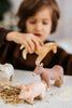 A child, blurred in the background and wearing a brown sweater, plays with a Handmade Wooden Pig toy alongside donkey and sheep figures. The toys rest on a table scattered with grains and straw, showcasing beautifully handcrafted pieces from Serbia.
