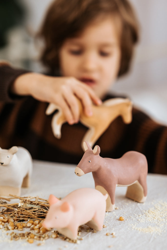 A child, blurred in the background and wearing a brown sweater, plays with a Handmade Wooden Pig toy alongside donkey and sheep figures. The toys rest on a table scattered with grains and straw, showcasing beautifully handcrafted pieces from Serbia.