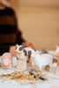 A Handmade Wooden Sheep figurine, crafted from linden wood, sits on a straw-scattered surface alongside farm animals. A person in a brown and black striped shirt is partially visible in the background.
