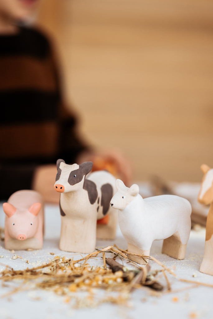 A Handmade Wooden Sheep figurine, crafted from linden wood, sits on a straw-scattered surface alongside farm animals. A person in a brown and black striped shirt is partially visible in the background.