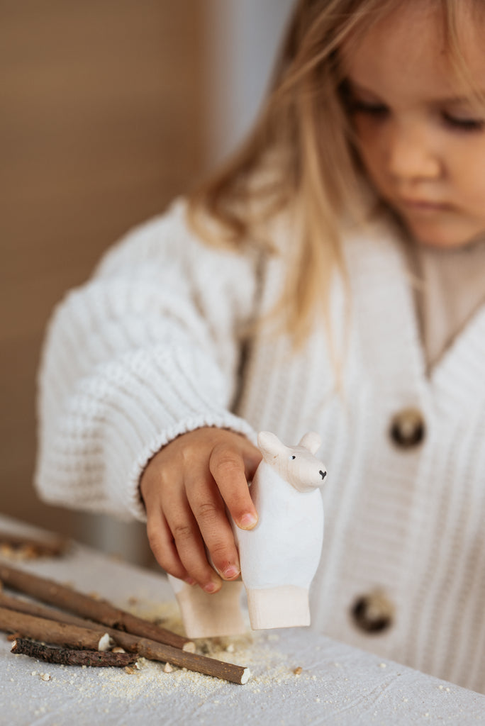 A child in a cozy sweater focuses on playing with a Handmade Wooden Sheep on a table, surrounded by scattered craft materials. The figurine and materials are coated with child-safe paint for imaginative fun.