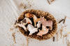 A woven basket filled with handcrafted linden wood farm animal figures, such as a cow, pig, goat, horse, and the Handmade Wooden Donkey painted with child-safe paint, rests on a fabric surface scattered with small twigs and grain.