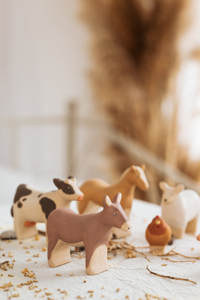 A collection of small, handcrafted linden wood farm animals like the Handmade Wooden Donkey, a black and white cow, a light brown horse, a white goat, and a red-brown hen with child-safe paint are displayed on a table scattered with seeds and grains.