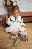 A young girl in a white dress with black trim plays on a carpeted floor with toy animals, including a zebra and the Handmade Wooden Hippo. In the background, a wicker basket and a flower vase enhance the charming scene with child-safe paint accents.