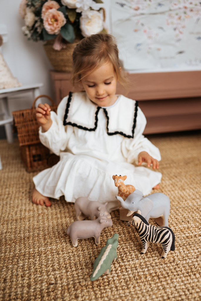 A young girl in a white dress with black trim plays on a carpeted floor with toy animals, including a zebra and the Handmade Wooden Hippo. In the background, a wicker basket and a flower vase enhance the charming scene with child-safe paint accents.