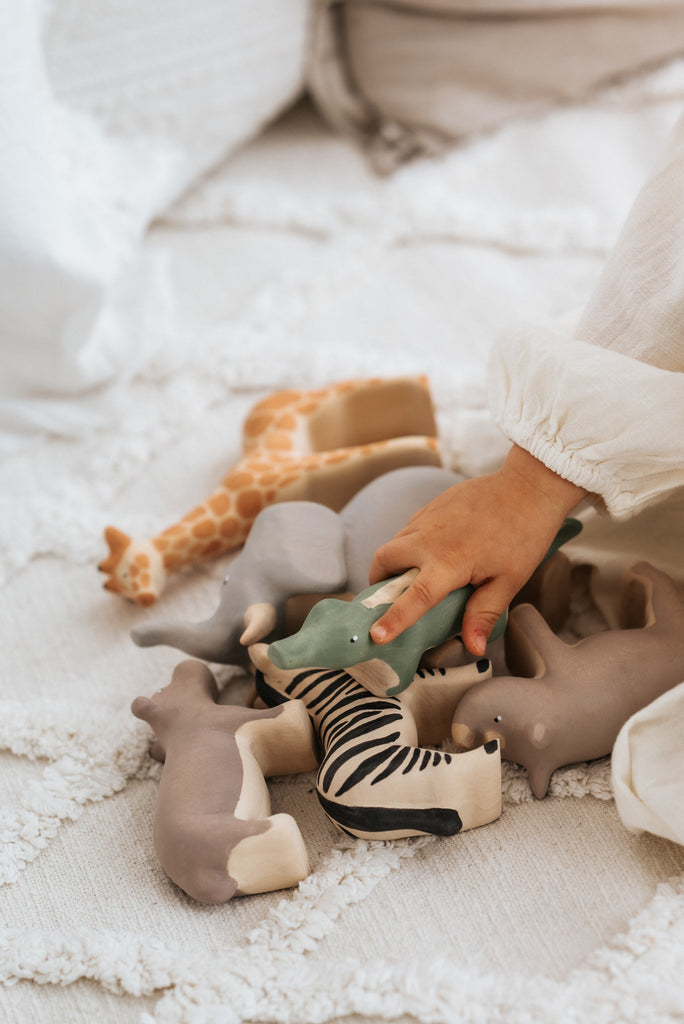 A child's hand reaches for toy animals like a giraffe, elephant, zebra, and rhinoceros on a cream blanket, including a Handmade Wooden Hippo finished with child-safe pastel paint.