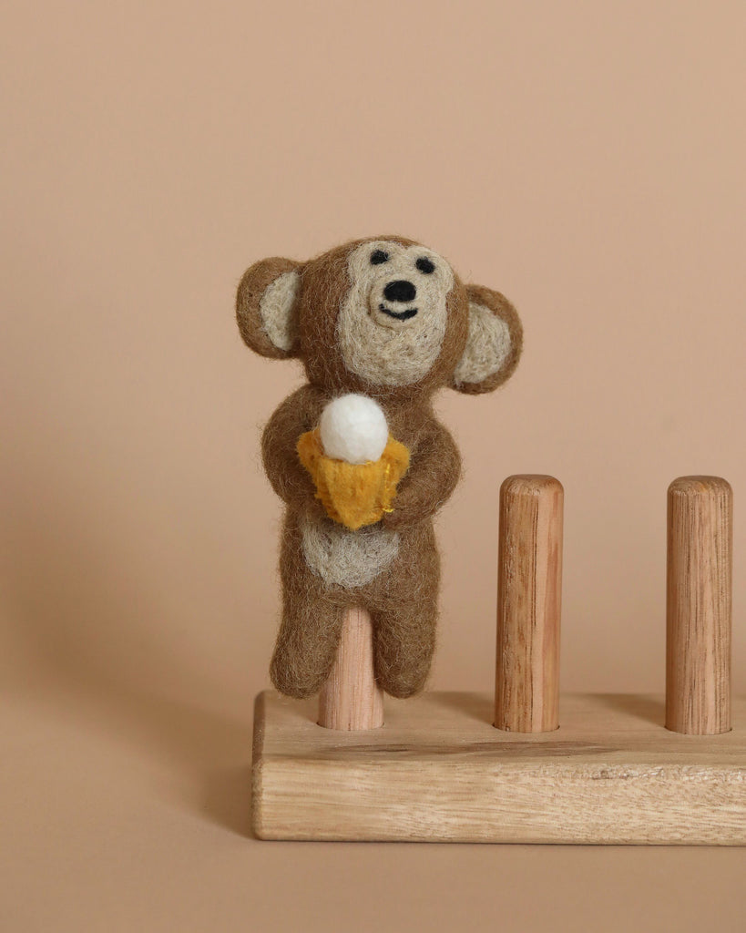 A small, brown needle felted monkey finger puppet is holding a miniature banana. It is perched on a wooden stand with three vertical pegs, against a beige background.