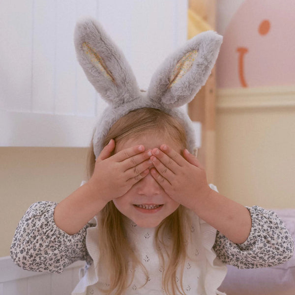 A young girl with long blonde hair playfully covers her eyes, wearing the Olli Ella Fluffle Bunny Headband - Smoke. Her white dress showcases a floral pattern in a softly lit room with pastel pink accents on the walls.