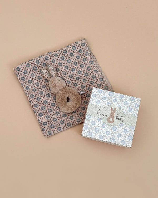 The Maileg Bunny Rattle Set - Cream Peach includes a plush bunny toy and a charming rattle on a small blue-beige blanket, alongside a floral gift box with "Bunny Baby" text, making it an ideal gift against the soft, neutral beige background.