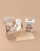 The Newborn Gift Set - Natural featuring a knitted baby blanket, Cuddle + Kind plush bear, socks, and bib sits beside an "Our Very Little Library" box on a beige background.