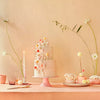 Elegant dessert table setup for a fairy garden party with a three-tiered cake adorned with delicate pastel flowers, surrounded by Meri Meri Mushroom Birthday Candles, flowers in vases, and bowls of sweets on a peach.
