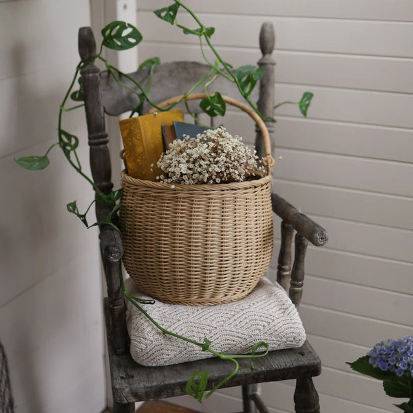 The Olli Ella Gathering Basket, filled with books and white flowers, sits on an antique wooden chair adorned with a green vine. A folded white blanket rests on the seat, creating a serene Easter ambiance against the backdrop of white paneled walls.