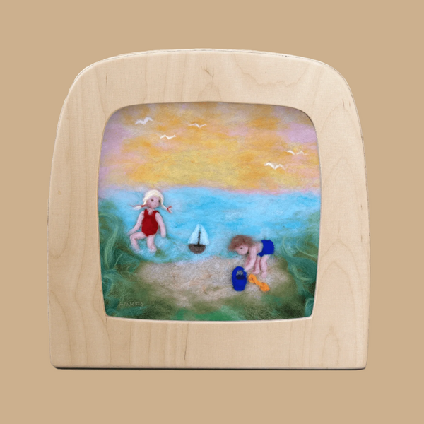 A charming wool felt silhouette for the Toverlux Lamp features two children playing on the beach with a toy boat, pail, and shovel beneath a yellow-blue sky with birds, bordered by grass—capturing the appeal of Magic Silhouettes.
