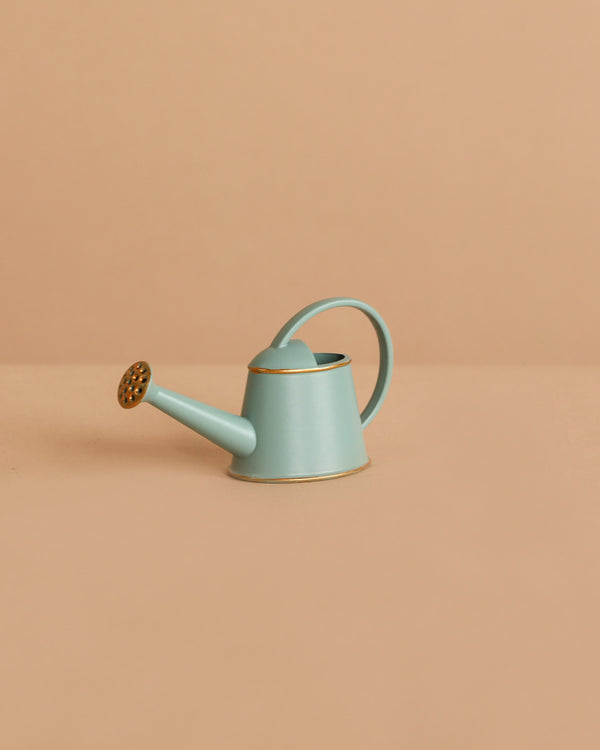 The Maileg Watering Can, Mouse - Light Blue, is a small metal accessory with a gold spout and trim, displayed on a beige surface and background—ideal for adding charm to any play scene.