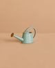The Maileg Watering Can, Mouse - Light Blue, is a small metal accessory with a gold spout and trim, displayed on a beige surface and background—ideal for adding charm to any play scene.