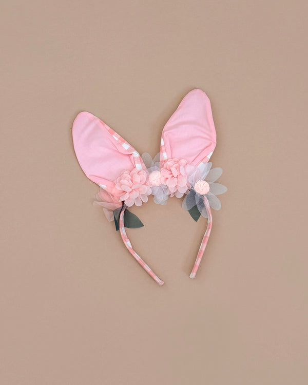 The Embellished Gingham Bunny Headband showcases pink fabric ears adorned with pastel pink and gray organza flowers and green leaves, set on a charming pink gingham band against a beige backdrop.