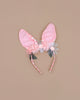 The Embellished Gingham Bunny Headband showcases pink fabric ears adorned with pastel pink and gray organza flowers and green leaves, set on a charming pink gingham band against a beige backdrop.