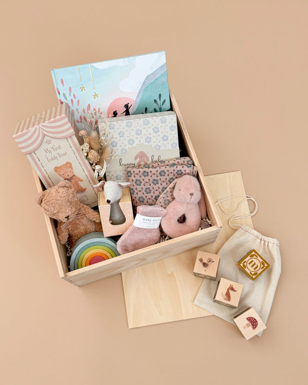 The Newborn Gift Set - Pink features a wooden box with baby items: teddy bear, socks, soft toy, Grimm's Small Wooden Rainbow, Uncle Goose Watercolor ABC Blocks, brush, and Let's Go Explore Book—all displayed on a beige background.