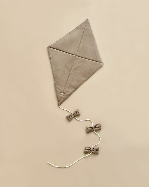 The Velvet Kite Wall Decor - Brown features a neutral-colored velvet kite with a simple triangular design, resting on a beige surface. It has a long string that ends in three small bows for both aerodynamics and decoration. Certified by OekoTex® Standard 100 and crafted from plain fabric, this minimalistic kite is ideal for decorating children's rooms.