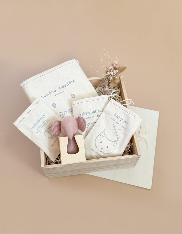 The Newborn Gift Set is a wooden box of newborn accessories, including a hooded swaddle, pilot hat, bib, and mittens made from organic cotton in elegant cream packaging. A charming small wooden toy completes this perfect baby gift against a neutral beige backdrop.