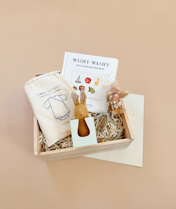 The Newborn Gift Set features a wooden box with a "Wishy Washy" book, an organic cotton baby romper in a drawstring bag, a wooden bunny toy, dried flowers, and crinkle paper filler, all beautifully displayed against a neutral background.