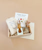 The Newborn Gift Set features a wooden box with a "Wishy Washy" book, an organic cotton baby romper in a drawstring bag, a wooden bunny toy, dried flowers, and crinkle paper filler, all beautifully displayed against a neutral background.