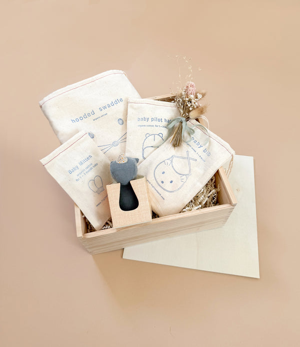 The Newborn Gift Set features a wooden box with a hooded swaddle, pilot hat, mittens, and toy—all made from organic cotton in neutral tones. A small bouquet of dried flowers adds charm, making it the ideal baby gift.