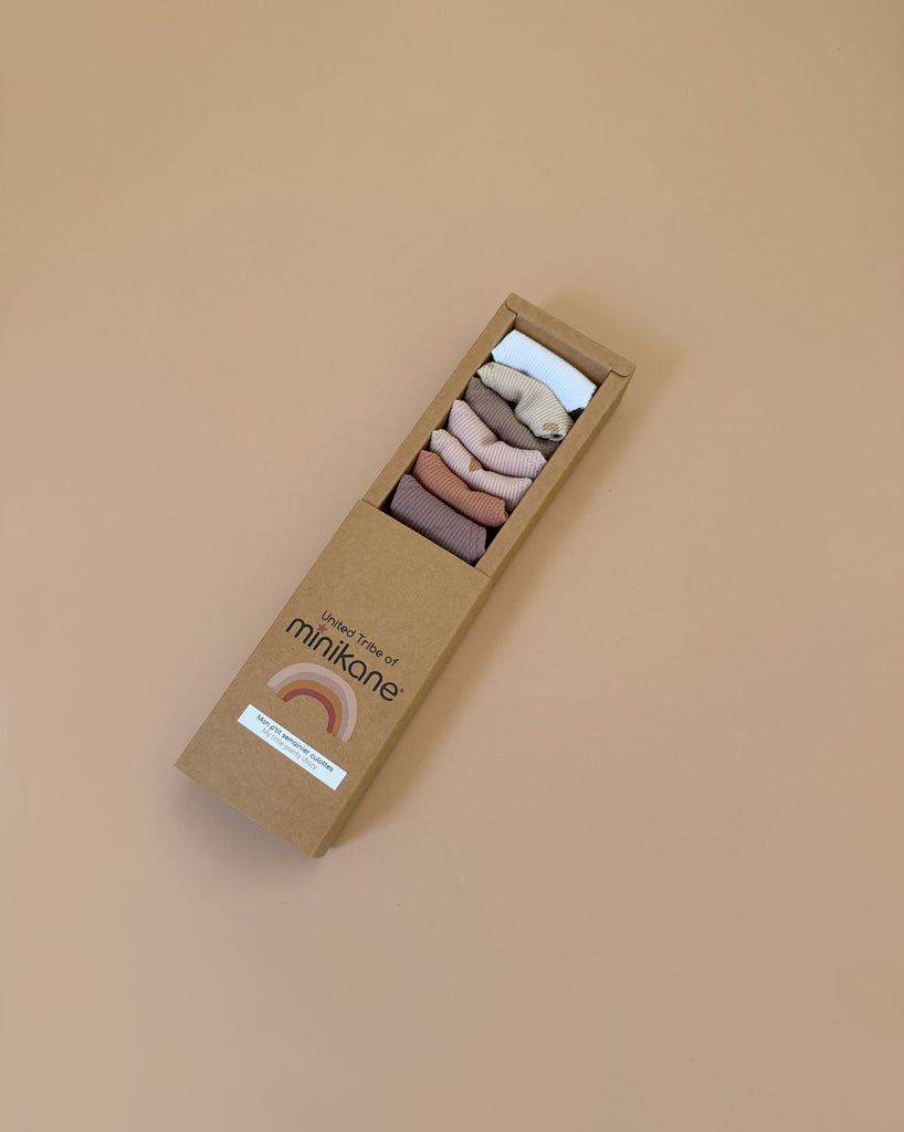 A Minikane "Little Hearts" Panties Weekly set for dolls, packaged in a rectangular cardboard box with a rainbow logo, holds six neatly folded, hand-washable ribbed panties. The muted shades of brown, beige, pink, and white are displayed in a gradient from dark to light. Perfect for Minikane Gordis dolls.