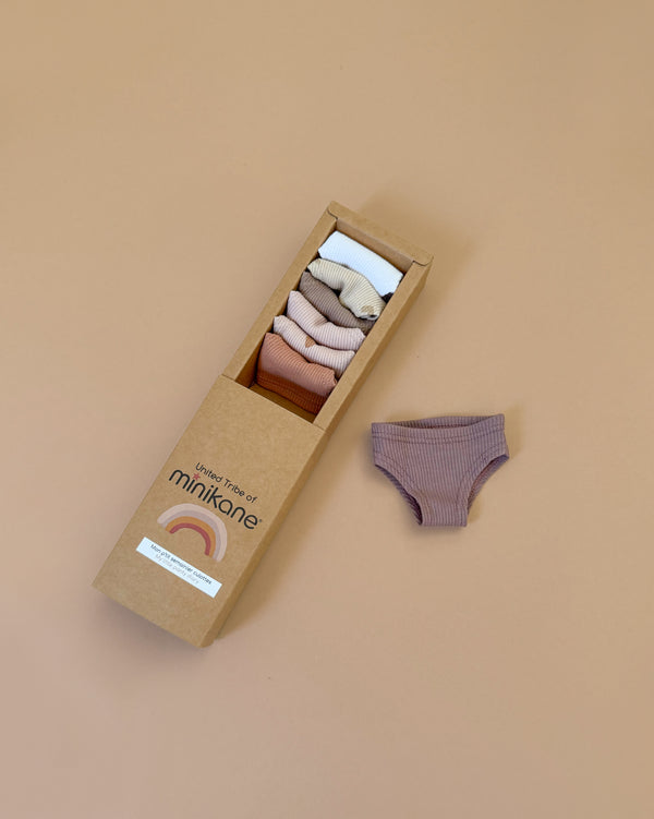 The Minikane kraft box contains the "Little Hearts" Panties Weekly set for dolls, featuring hand-washable fabric underwear in beige, pink, brown, and mauve. Perfectly designed for Minikane Gordis dolls and made in France, one mauve pair is displayed beside the box on a neutral beige background.