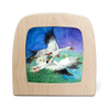 A child's illustration on a wooden frame, crafted with food-safe materials, shows two flying geese carrying a person in colorful garments. Stars trail behind, creating Magic Silhouettes against a shifting blue-to-green background. Product: Silhouette Only - George and the Snow Geese.
