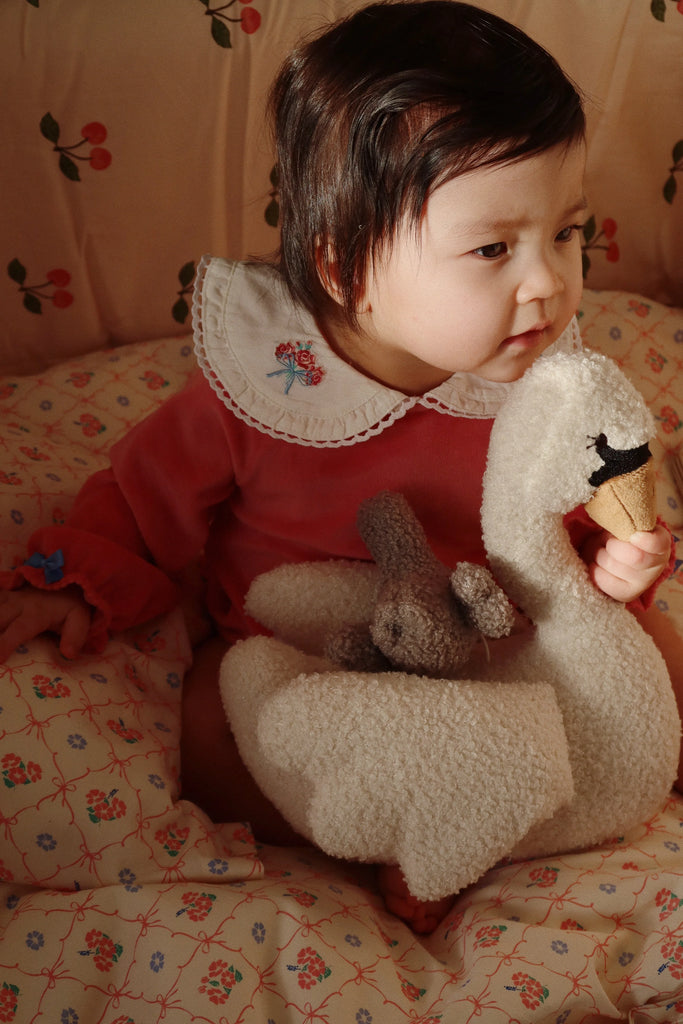 A baby with dark hair sits on a patterned blanket, clad in a pink outfit with a white ruffled collar, holding the Konges Sloejd Activity Music Swan—a plush toy that plays Brahms' Lullaby—and a small brown stuffed animal, all set against a background featuring floral patterns.