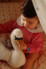A baby in a pink outfit sits on a floral-patterned bed, gently holding the Konges Sloejd Activity Music Swan. The scene is softly lit, creating a warm and cozy atmosphere reminiscent of a soothing melody.