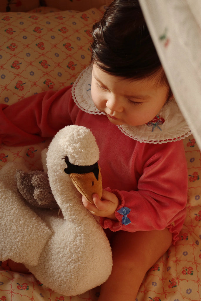 A baby in a pink outfit sits on a floral-patterned bed, gently holding the Konges Sloejd Activity Music Swan. The scene is softly lit, creating a warm and cozy atmosphere reminiscent of a soothing melody.