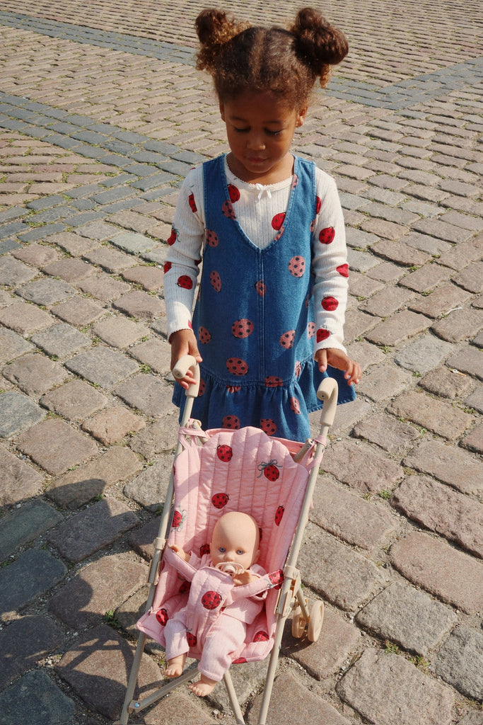 A child in a denim dress with ladybug patterns and a white long-sleeve shirt pushes a Konges Sloejd Doll Stroller - Ladybug, featuring double wheels, along the cobblestone path.