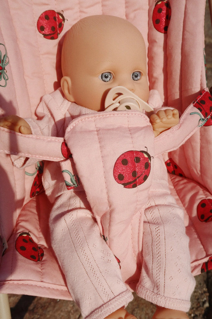 The Konges Sloejd Doll Stroller - Ladybug features a baby doll with blue eyes seated in an easy-to-fold pink stroller adorned with ladybug designs. The doll is dressed in a matching pink outfit, holds a pacifier, and enjoys stability for playful adventures in the sunlight.