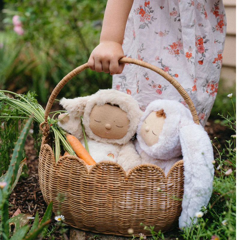 In a sunlit garden, a child with a floral dress holds her Olli Ella Scalloped Petal Basket - Natural, filled with two fluffy stuffed animals and a fresh carrot. Surrounded by greenery, it's the ideal springtime adventure setting.
