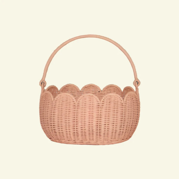 The Olli Ella Petal Basket in Seashell Pink features a scalloped design with a sturdy arched handle, crafted from natural wicker for rustic charm. Perfect for spring adventures or displaying against a neutral backdrop, it seamlessly combines practicality and style.